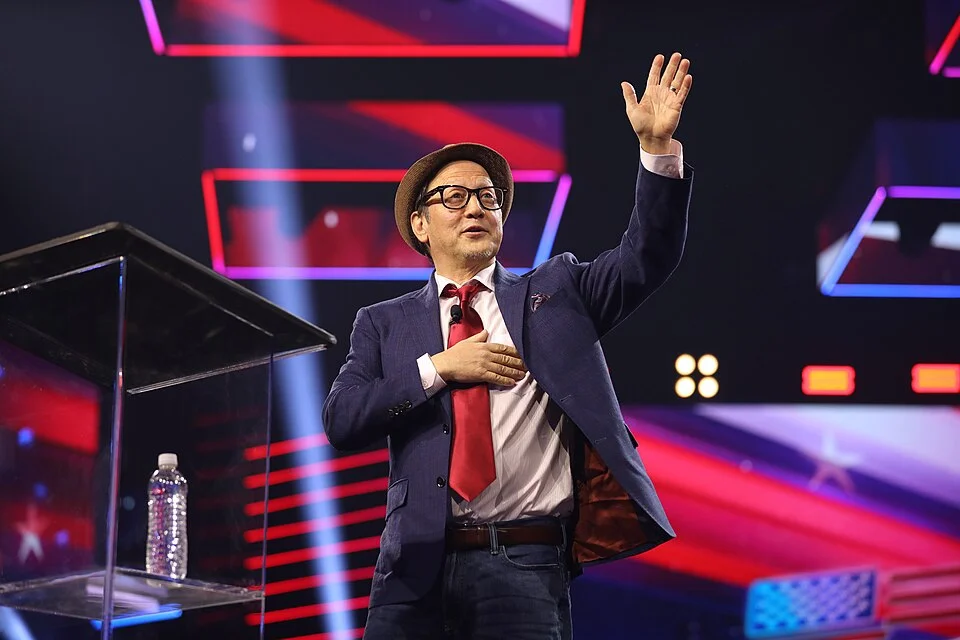 Rob Schneider on stage with hand on heart during a live appearance