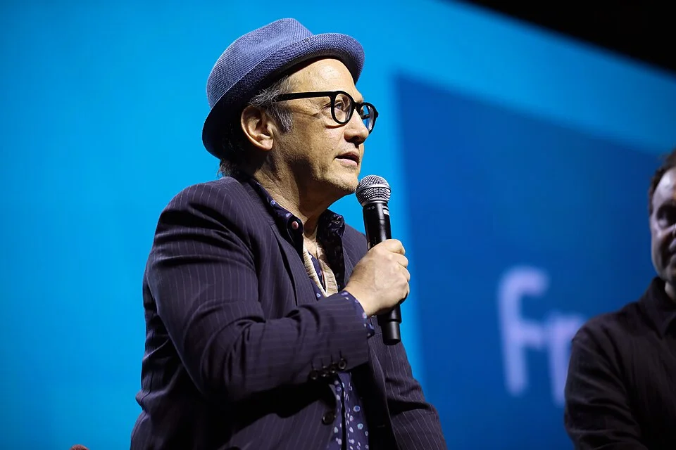 Rob Schneider speaking into a microphone in a hat and glasses at a live event