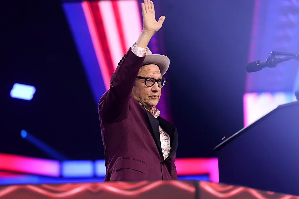 Rob Schneider waving to the crowd in a maroon suit and hat on stage