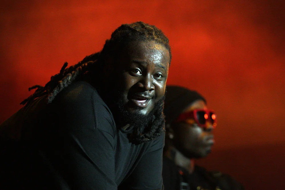 T-Pain smiling in a black shirt against a red backdrop