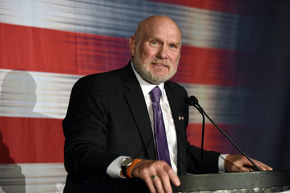 Terry Bradshaw speaking at a podium in front of an American flag mantlemagazine.com