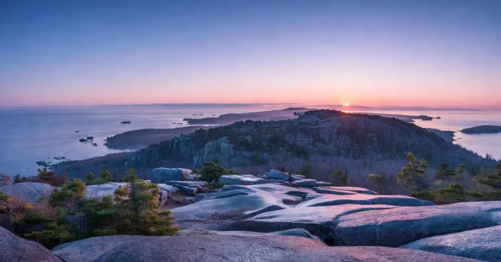 Panoramic sunrise view from Cadillac Mountain summit showing granite rocks, coastline, and islands in Atlantic Ocean