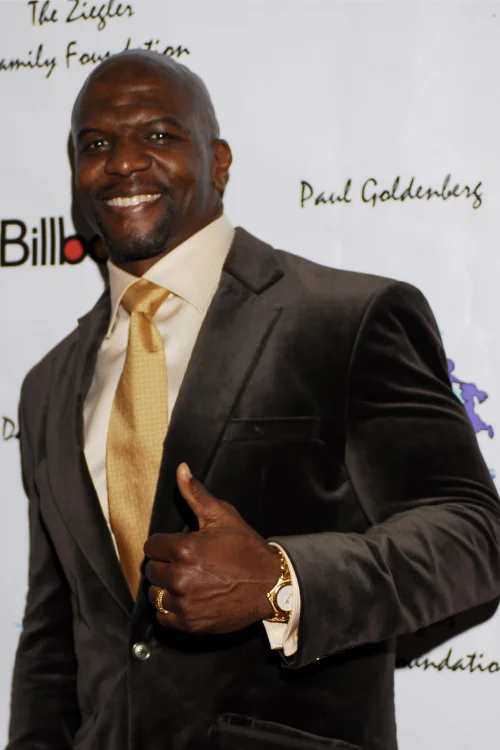 Terry Crews smiling and giving a thumbs-up in a dark suit and gold tie at a red carpet event
