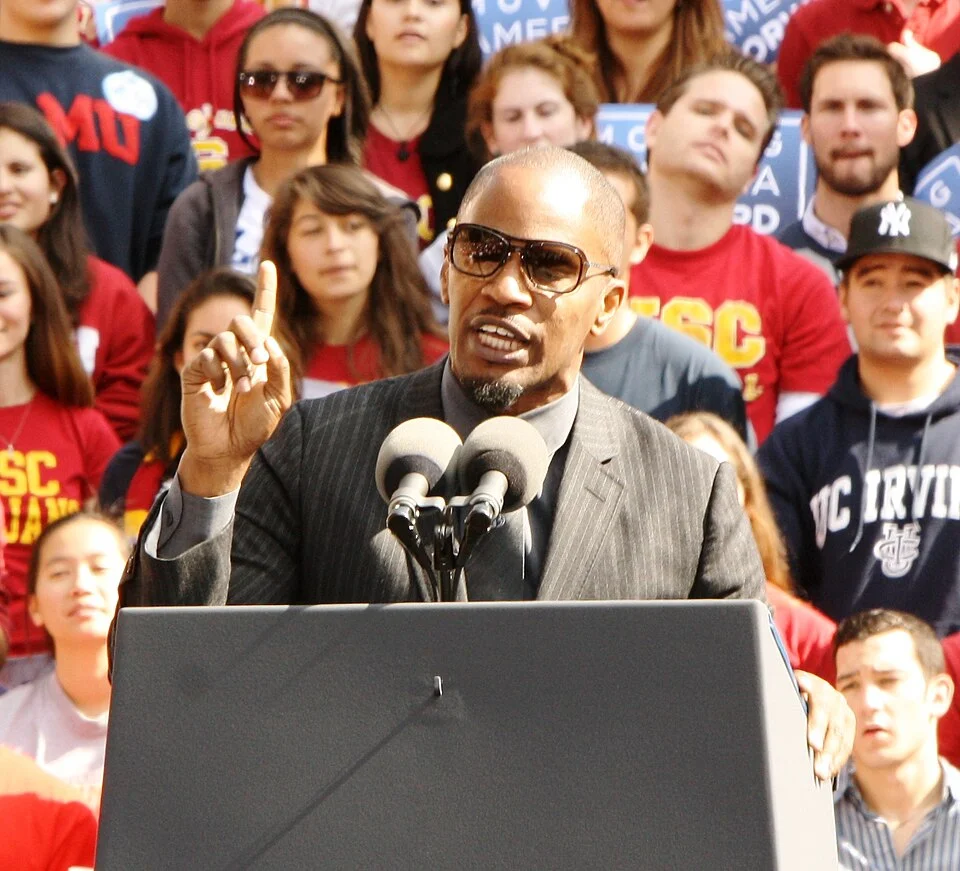 "Jamie Foxx speaking at a podium wearing sunglasses and pinstripe suit addressing a crowd of students and supporters"