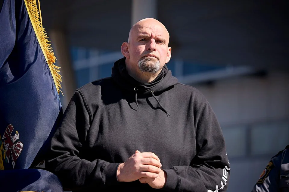 John Fetterman at an outdoor event beside the Pennsylvania state flag — mantlemagazine.com