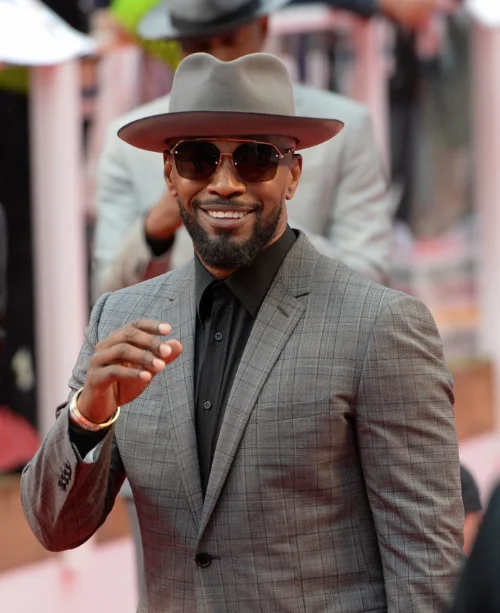"Jamie Foxx smiling in checked gray suit jacket, black shirt, and wide-brim hat at red carpet premiere"