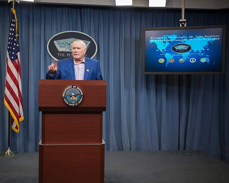 Terry Bradshaw speaking at a Pentagon podium, with a screen behind him welcoming the 4x Super Bowl champion and 2x Super Bowl MVP