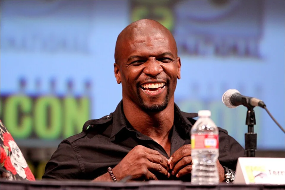 Terry Crews laughing at a Comic-Con panel, seated behind a microphone with the convention backdrop visible