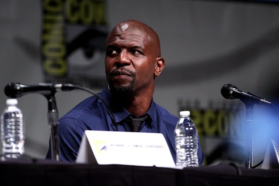 "Terry Crews speaking at Comic-Con panel discussion, wearing blue shirt and black tie, seated at table with microphones and water bottles"