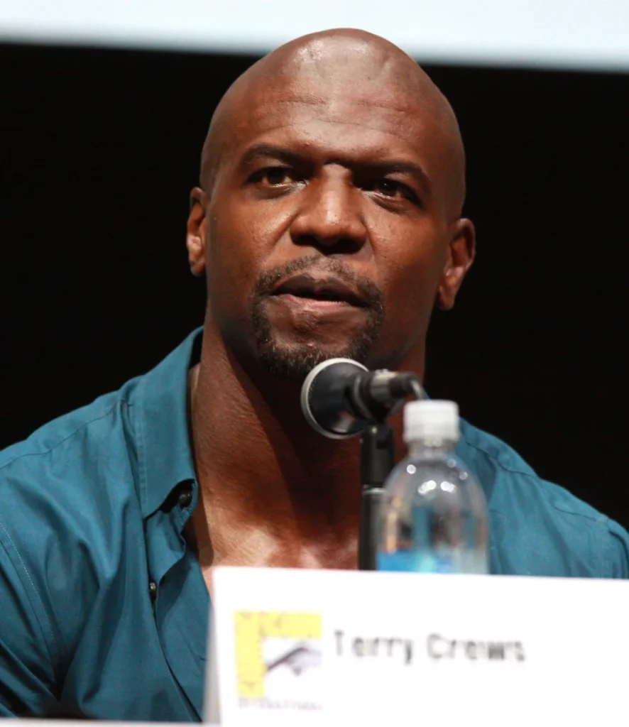 Terry Crews speaking into a microphone at a Comic-Con panel, with his name card visible on the table