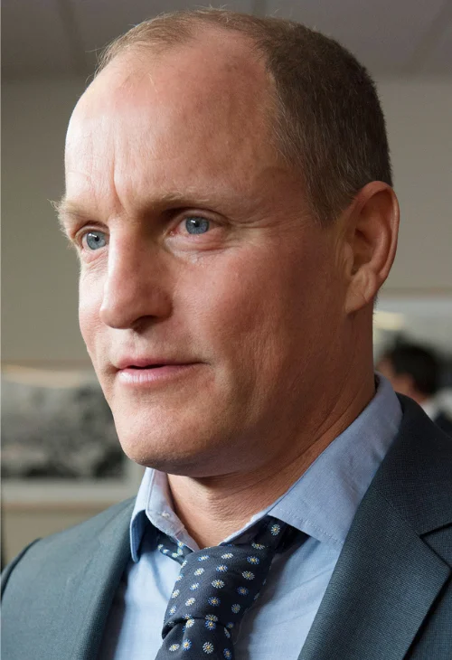 Close-up portrait of Woody Harrelson in a grey suit, light blue shirt and patterned tie, looking off camera