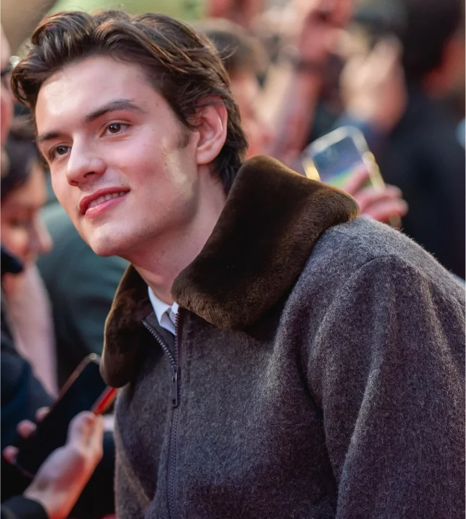 Louis Partridge smiling in a fur-collared gray coat as fans hold up phones behind him