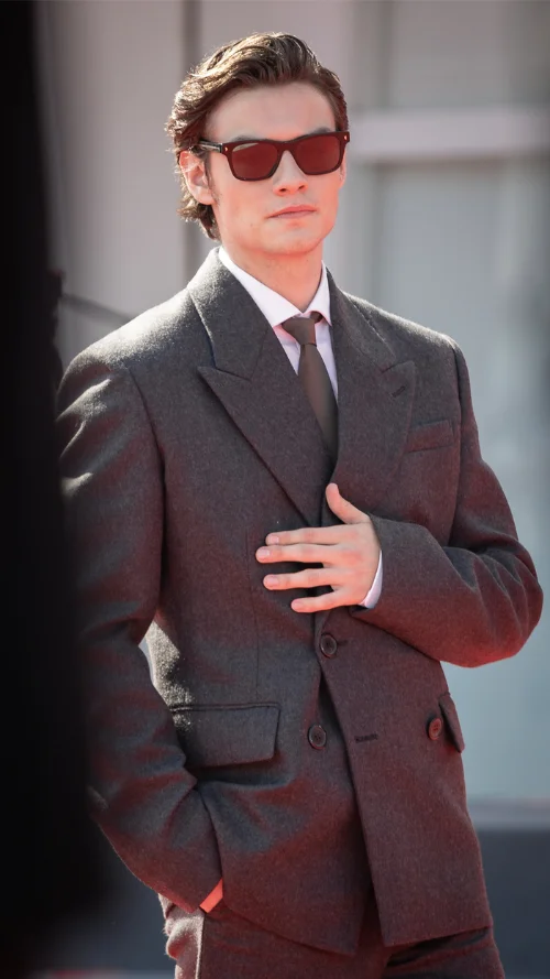 Louis Partridge posing in a gray suit and red-tinted sunglasses