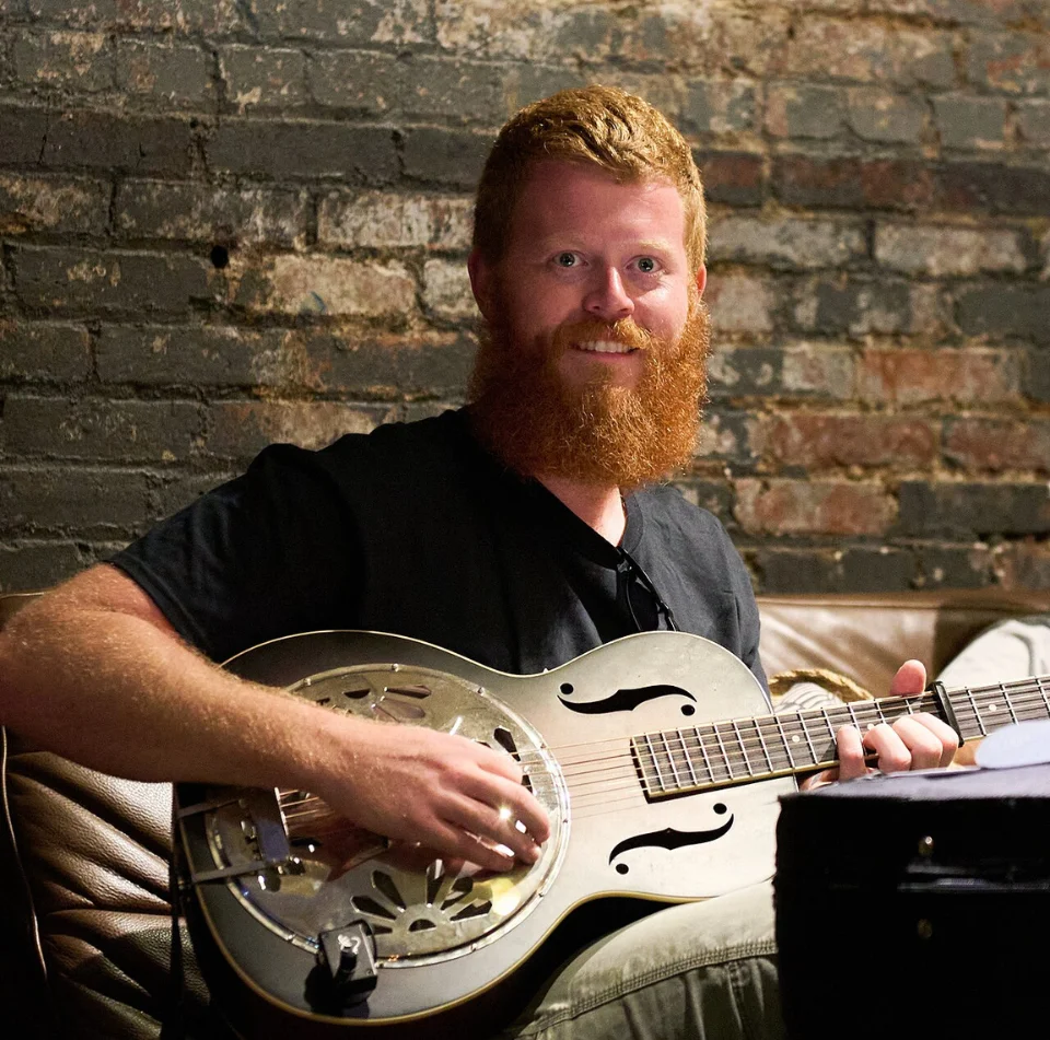 Oliver Anthony smiling with a resonator guitar against a brick wall