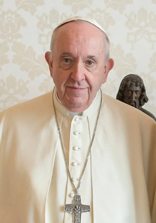 Pope Francis in white papal vestments and silver pectoral cross standing before a statue of Jesus Christ