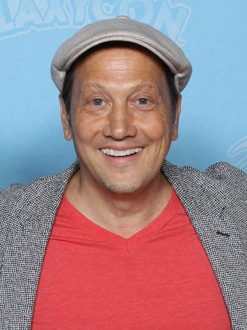 Rob Schneider smiling while speaking at a microphone during a public appearance, wearing casual attire