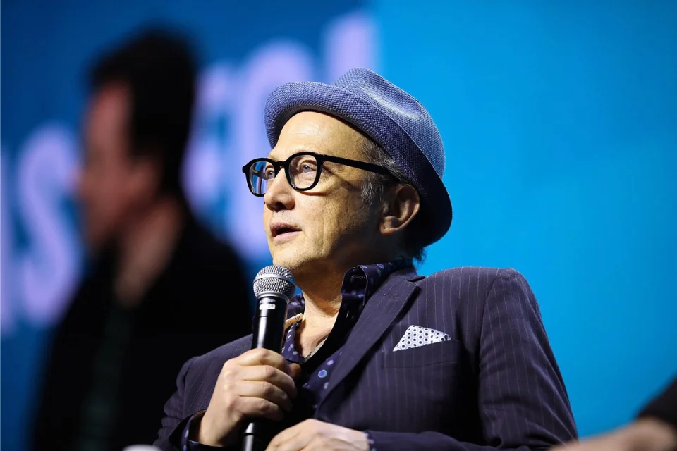 Close-up of Rob Schneider holding a microphone on a blue-lit stage