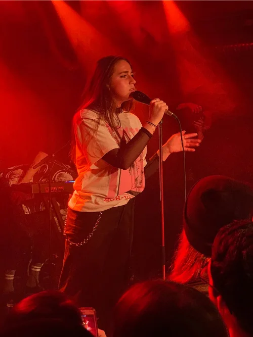 A young Tate McRae singing into a microphone at an early intimate venue show under red stage lighting