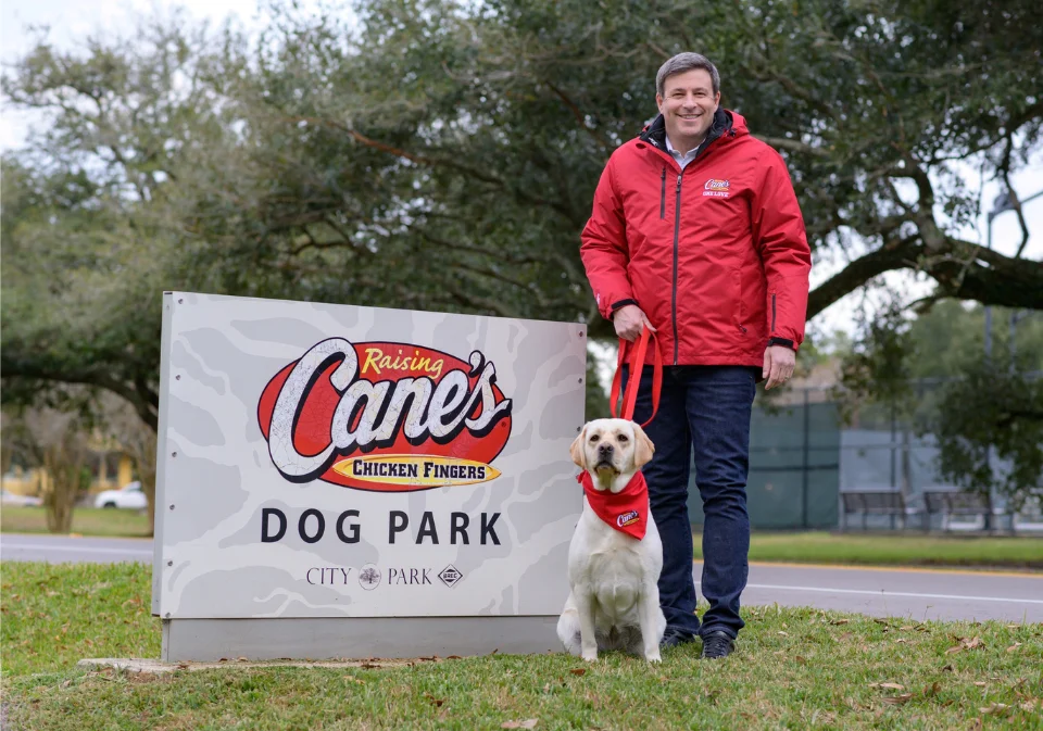 Todd Graves, founder of Raising Cane's, standing with a yellow Labrador holding a chicken finger next to a Raising Cane's Dog Park sign